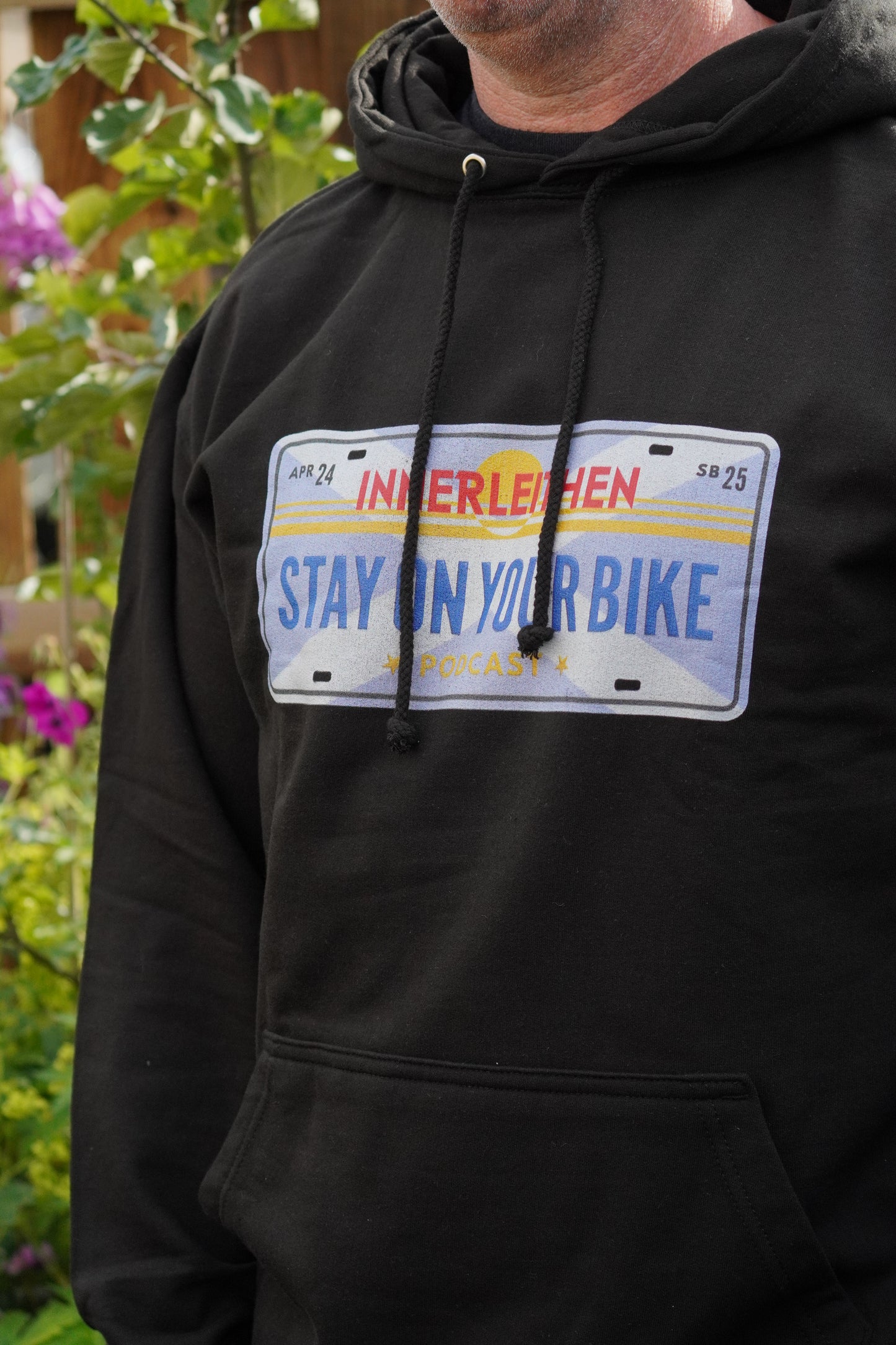 Close-up of the Californian style licence plate design on black Loam Days Hoodie, worn by Nigel Page, featuring 'Stay on Your Bike' and 'Innerleithen' text with horizon graphic – mountain biking merch and souvenir from Tweed Valley, inspired by Stay on Your Bike podcast
