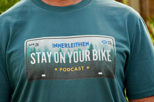 Close up of the 'Stay On Your Bike' , worn by Nigel Page and 'Innerleithen' print on the Into The Woods T-shirt, blue organic cotton, short sleeve, podcast merch and mtb souvenir form the Tweed Valley. 