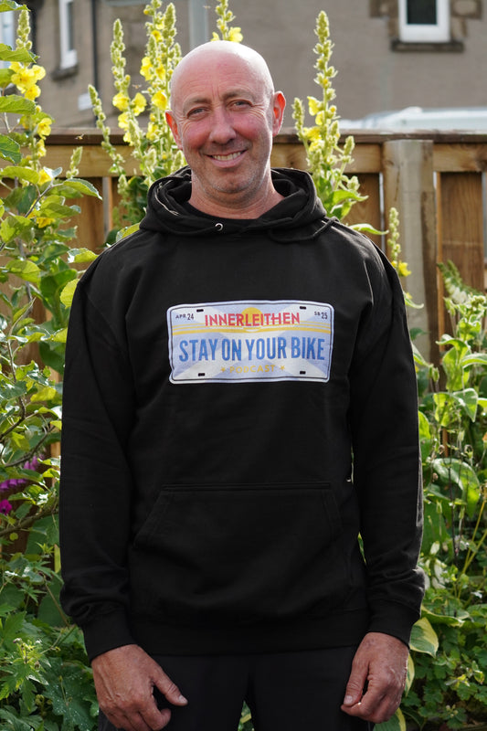 Nigel Page wears Black unisex Loam Days hoodie jumper made from cotton blend, featuring a Californian style licence plate print with 'Stay on Your Bike' and 'Innerleithen' text and horizon design – mountain biking merch and souvenir from Innerleithen in the Tweed Valley